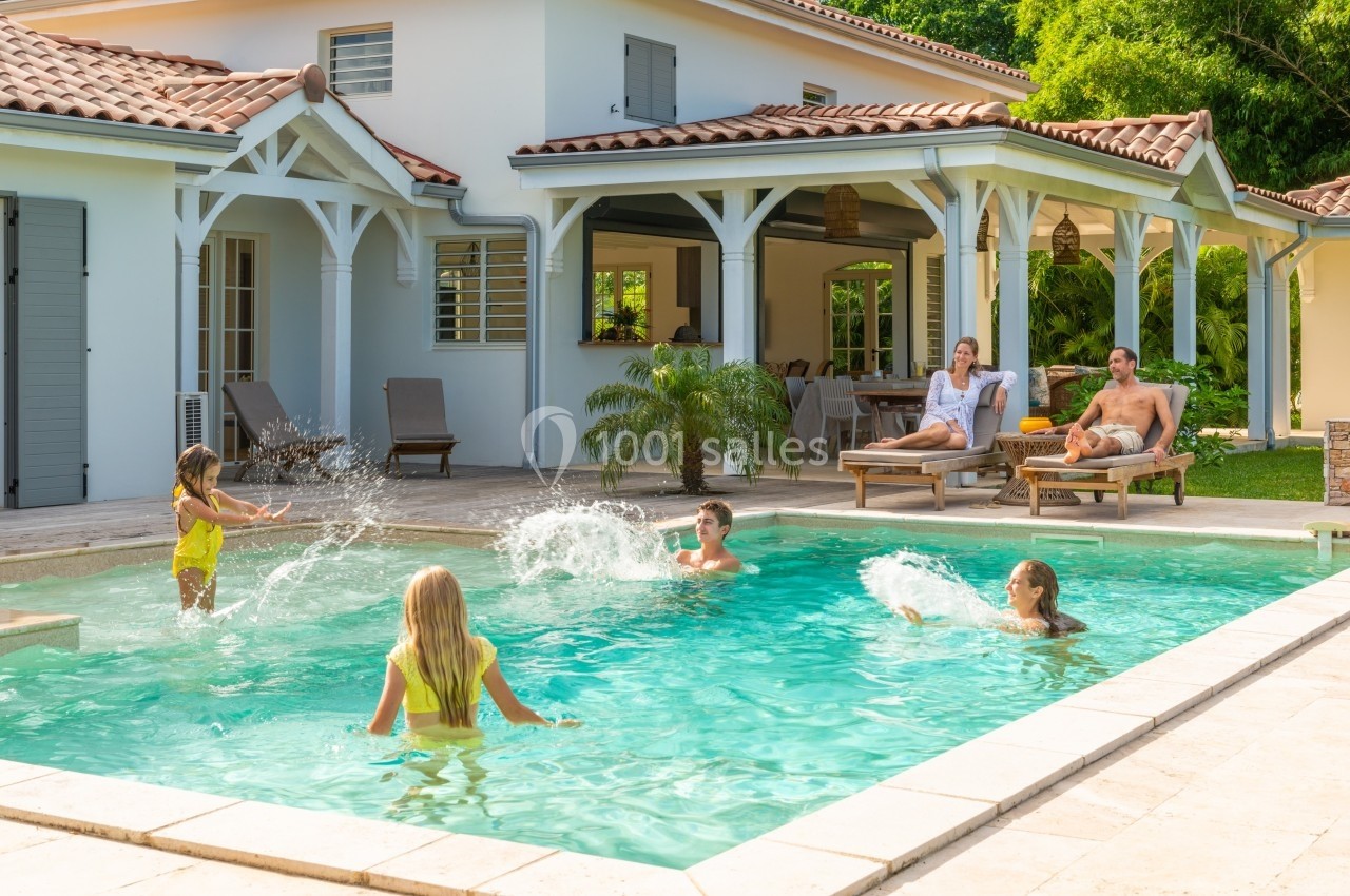 Des personnes profitent d'une piscine dans le jardin d'une maison, entourées de végétation et de mobilier extérieur.