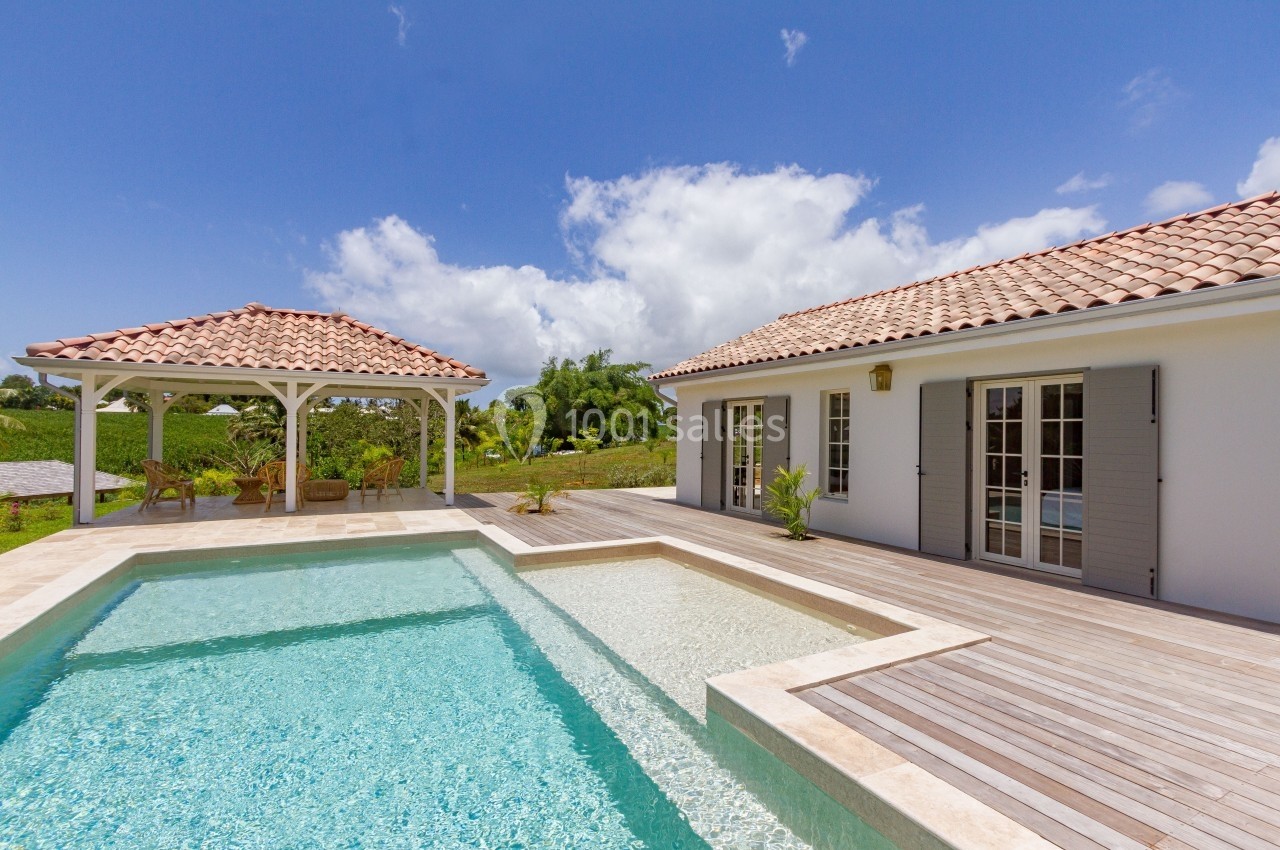Maison avec terrasse en bois, piscine et pergola, entourée de verdure sous un ciel bleu.