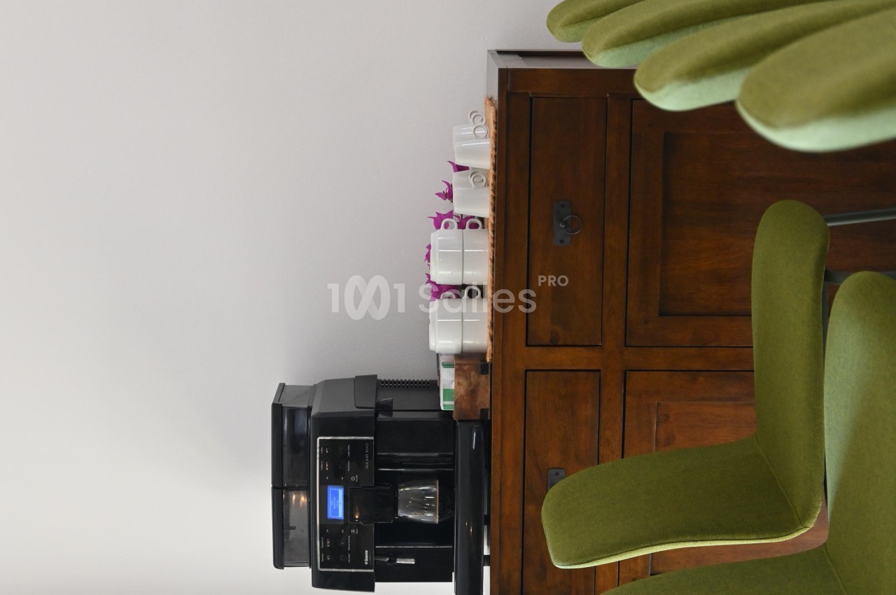 Machine à café noire posée sur un meuble en bois, entourée de tasses blanches et de chaises vertes.