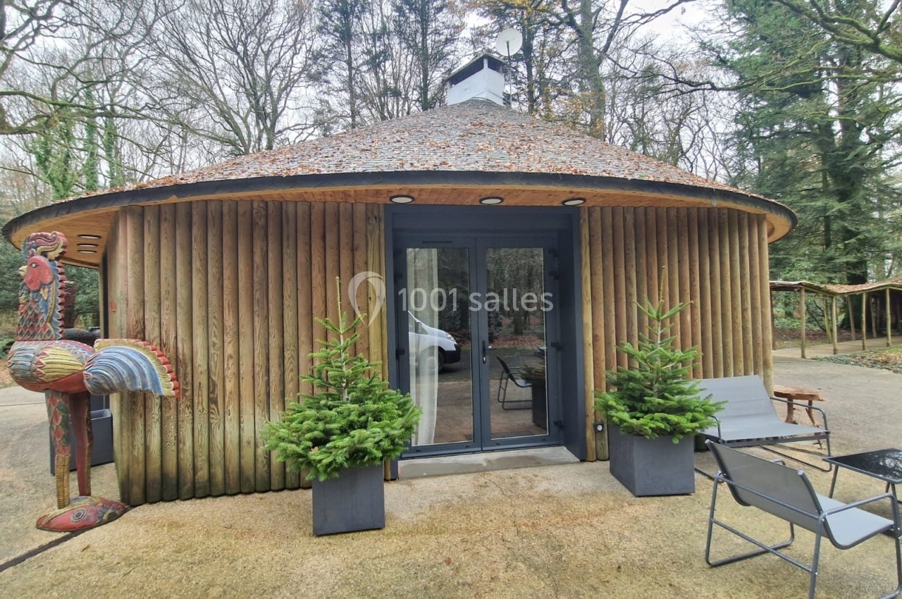 Cabane ronde en bois avec toit en ardoise, entourée d'arbres, deux chaises et plantes en pots devant l'entrée.