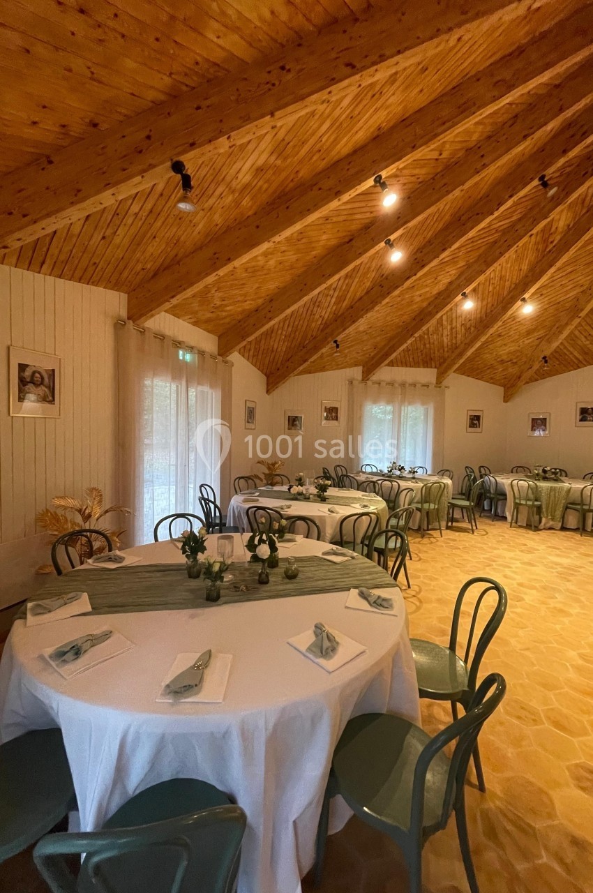 Salle décorée avec des tables rondes, nappes blanches et chaises vertes, sous un plafond en bois avec éclairage chaleureux.