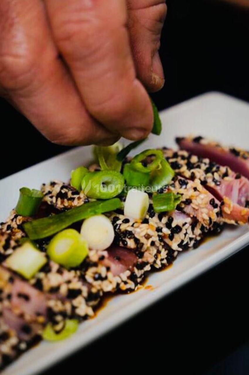 Main saupoudrant des rondelles d'oignons verts sur un plat de thon en croûte de sésame, présenté sur une assiette blanche.