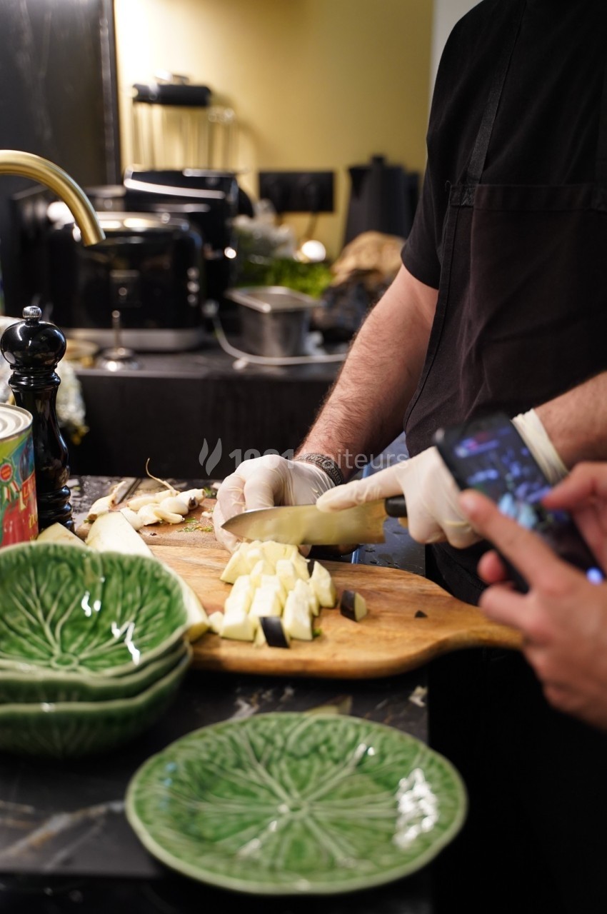 Un cuisinier découpe des légumes sur une planche en bois, avec des assiettes vertes en forme de feuille au premier plan.