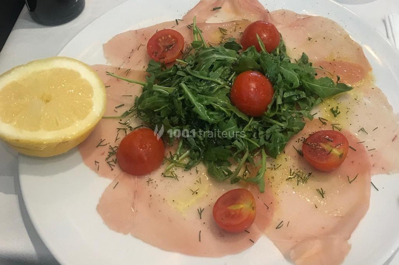 Assiette de carpaccio de poisson garnie de roquette, tomates cerises et herbes, accompagnée d'un demi-citron.