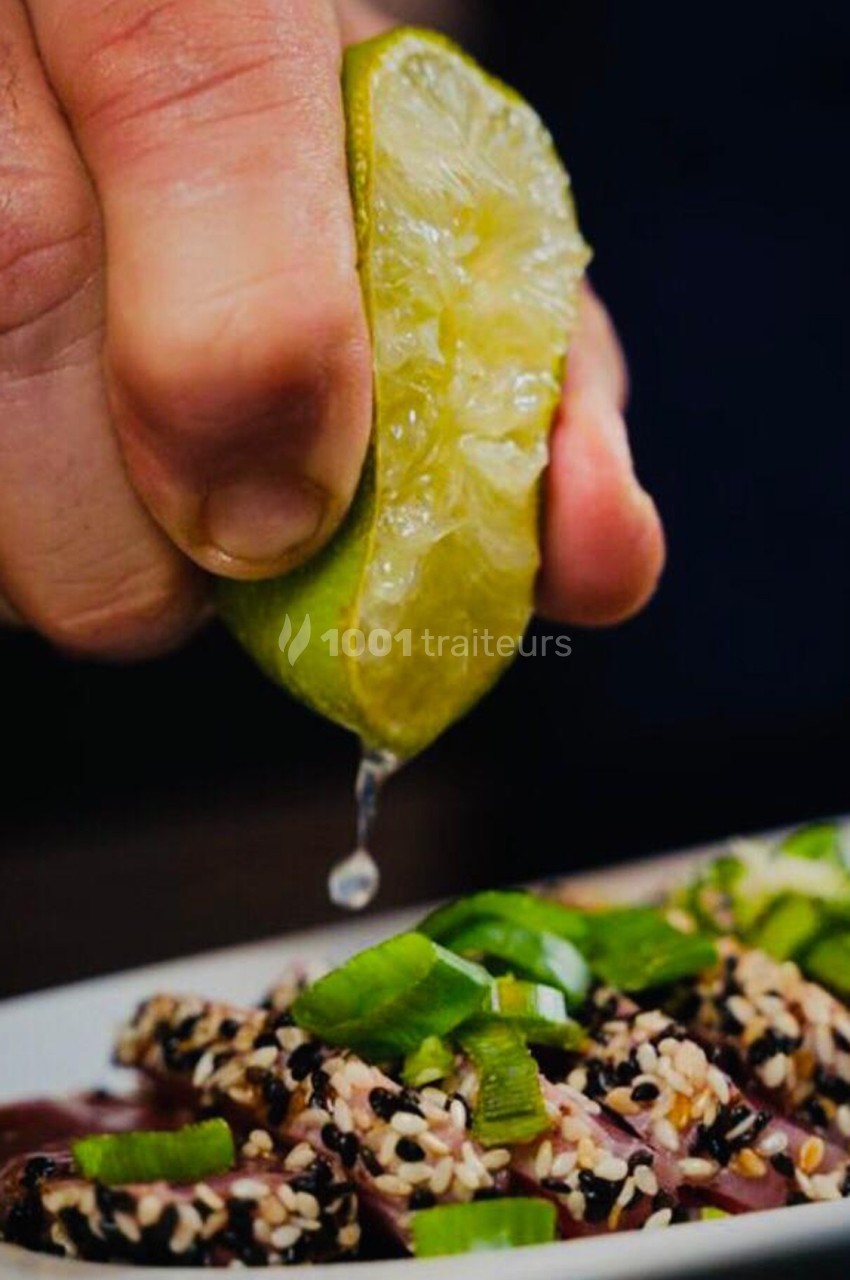 Une main presse un demi-citron vert au-dessus d'un plat garni de graines de sésame et de morceaux d'oignons verts.