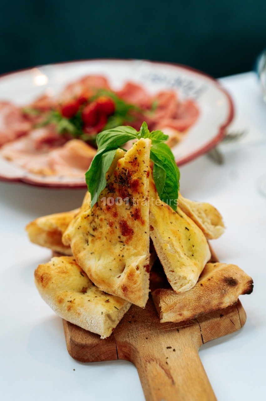 Tranches de pain plat doré garnies d'herbes, servies sur une planche en bois avec un plat de charcuterie en arrière-plan.