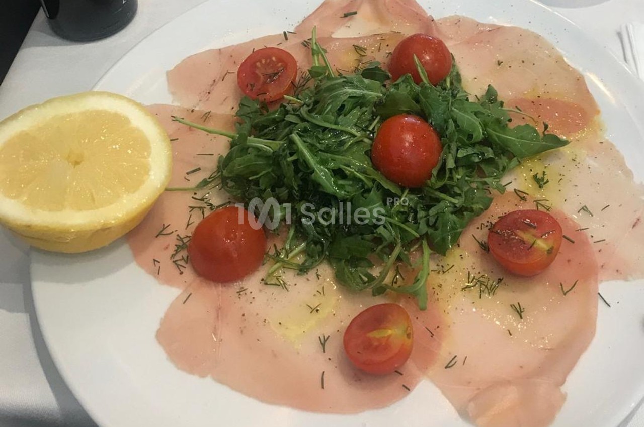Assiette de carpaccio de poisson garnie de roquette, tomates cerises et herbes, accompagnée d’un demi-citron.