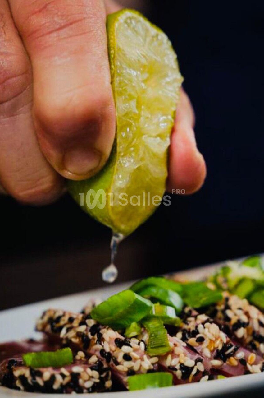 Une main presse un demi-citron vert au-dessus d'un plat garni de graines de sésame et de morceaux de légumes verts.