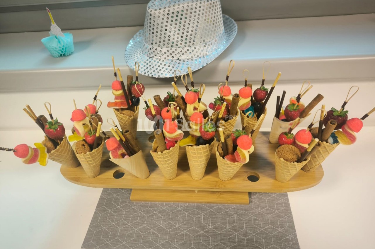 Présentoir en bois avec des cornets garnis de fruits, bonbons et biscuits, posé sur une table près d'un chapeau argenté.