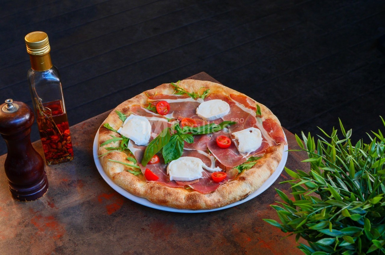 Pizza garnie de jambon cru, fromage, tomates cerises et roquette, servie sur une table avec huile pimentée et plante…