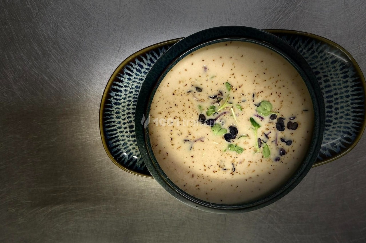 Bol de soupe crémeuse garnie de pousses vertes et de graines, posé sur une assiette sur une surface métallique.