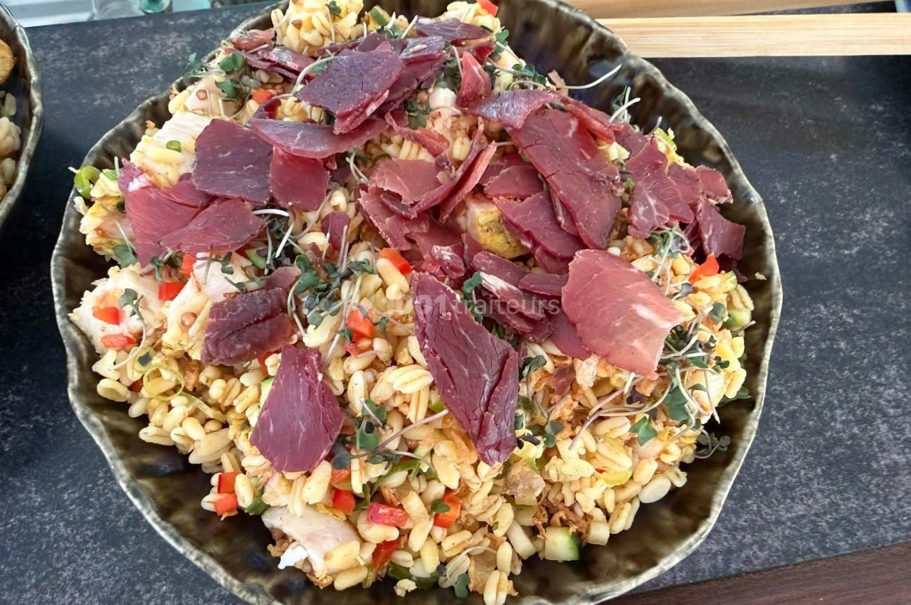 Assiette de salade de céréales garnie de tranches de viande séchée, légumes colorés et pousses fraîches.