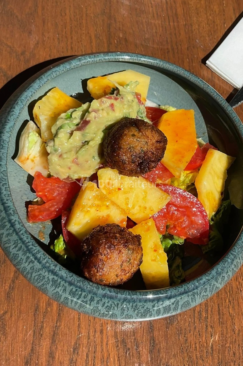 Assiette contenant une salade colorée avec tomates, ananas, guacamole et boulettes végétariennes sur une table en bois.
