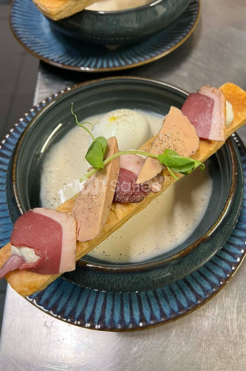 Assiette contenant une soupe crémeuse, garnie d'une pâte feuilletée avec foie gras, magret fumé et pousses de pois.