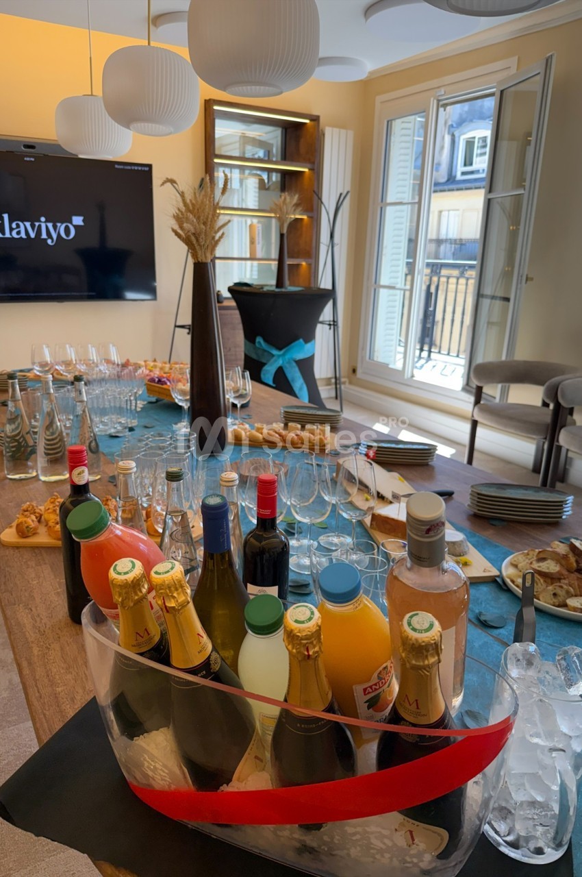 Table dressée avec boissons, verres, viennoiseries et amuse-bouches dans une salle lumineuse avec écran et fenêtres ouvertes.