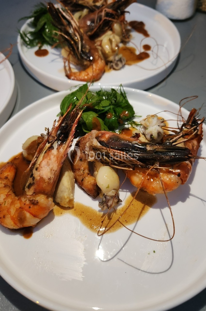 Assiette de crevettes grillées avec légumes verts, pommes de terre et sauce, servie sur une table grise.