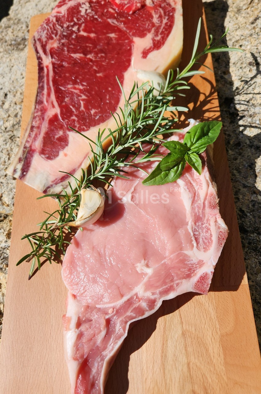 Deux morceaux de viande crue, l'un rouge et l'autre rose, posés sur une planche en bois avec des herbes aromatiques.
