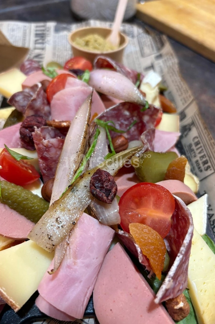 Plateau de charcuterie et fromages variés avec cornichons, tomates cerises, fruits secs et une sauce en arrière-plan.