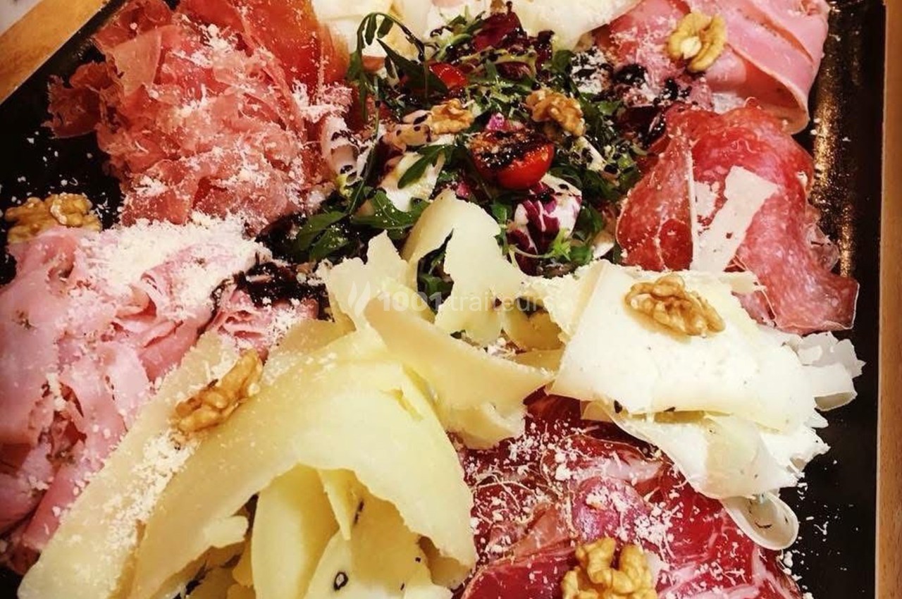 Plateau de charcuterie et fromages avec noix, herbes fraîches et copeaux de parmesan, présenté sur un plat noir.