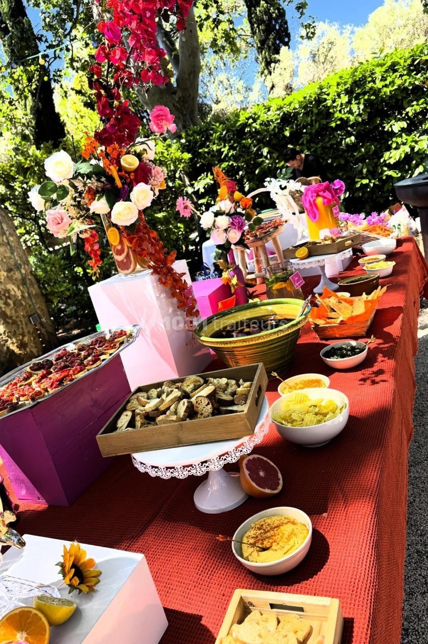 Buffet coloré en extérieur avec des plats variés, des fleurs décoratives et une nappe rouge.
