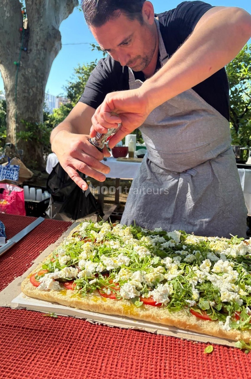 Un homme en tablier ajoute des herbes fraîches sur une grande tarte salée dans un jardin ensoleillé.
