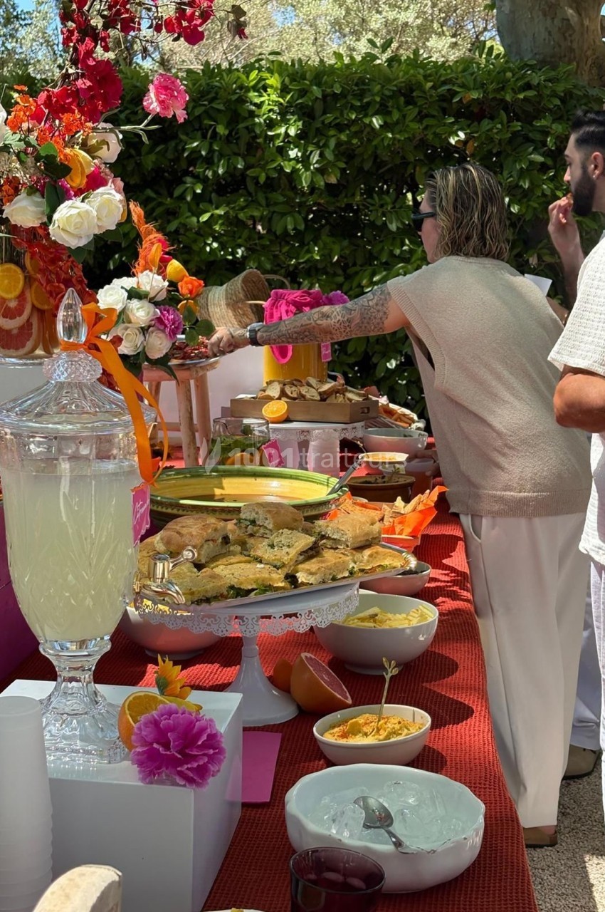 Buffet en extérieur avec diverses préparations culinaires, fleurs décoratives et boissons disposées sur une table rouge.