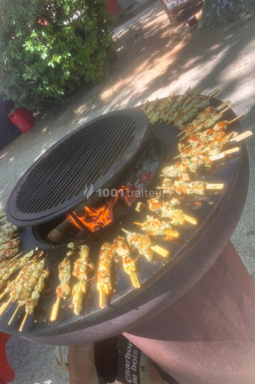 Brochettes de viande et légumes grillant sur un barbecue circulaire avec feu visible au centre.