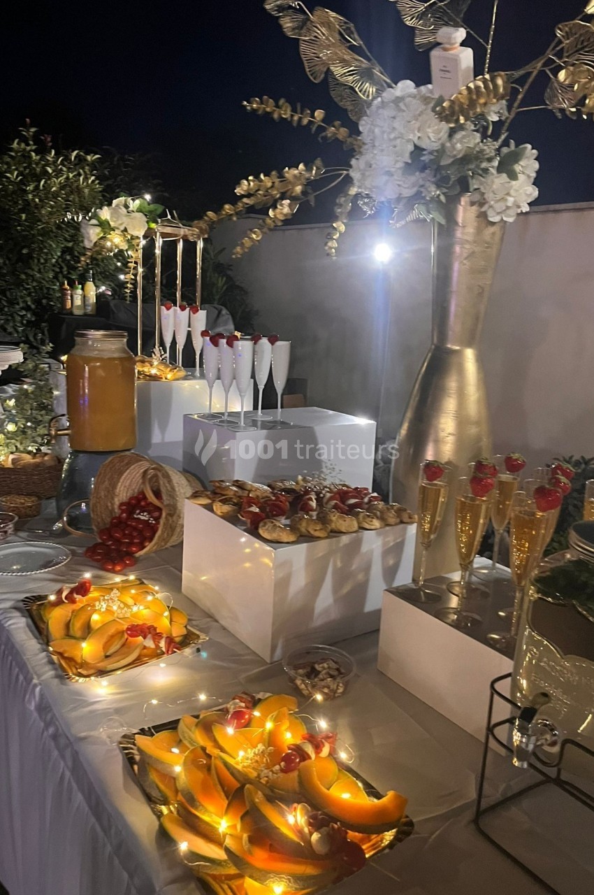 Buffet nocturne décoré avec des fruits, pâtisseries, boissons et guirlandes lumineuses, entouré de végétation.