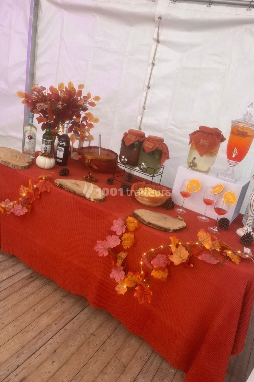 Table décorée pour l'automne avec boissons, fruits, fleurs, feuilles colorées et accessoires sur une nappe rouge.