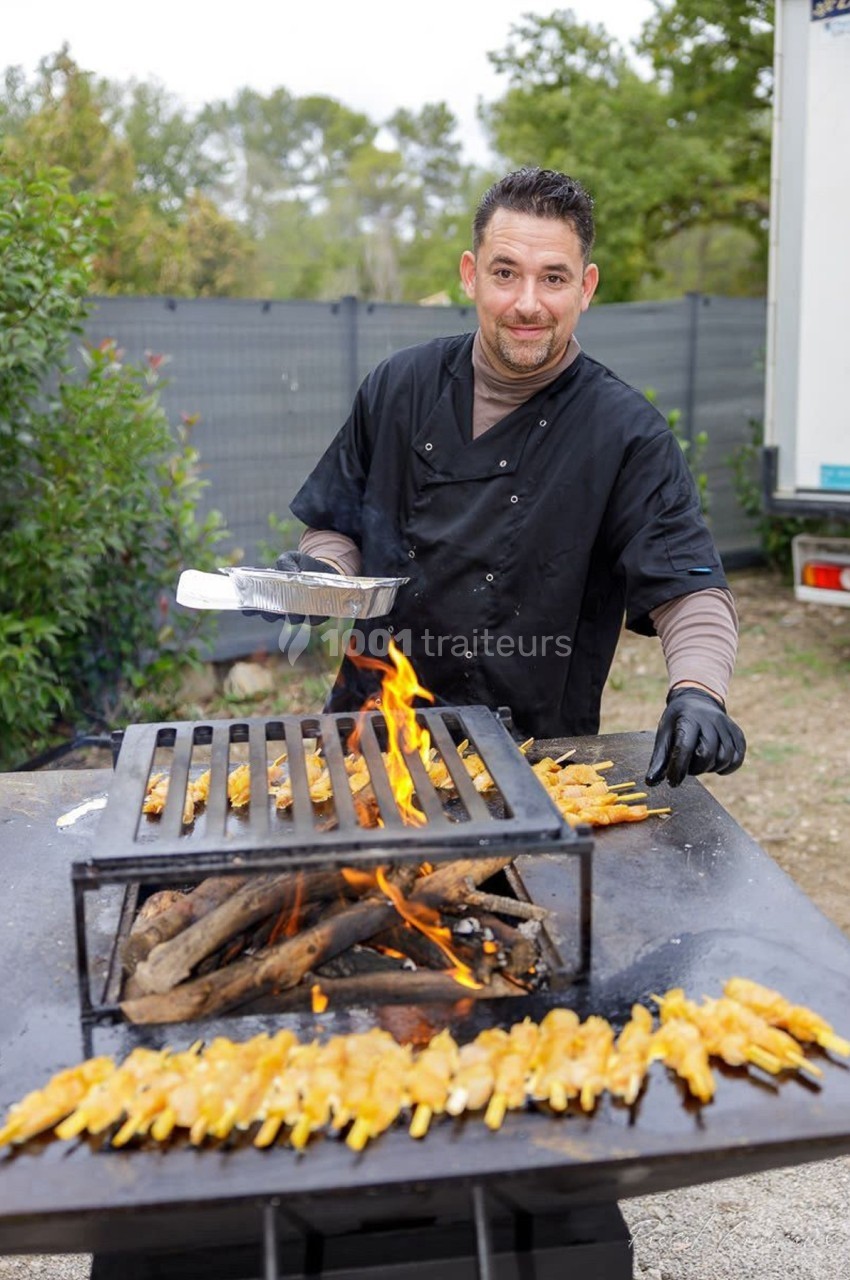 Un cuisinier prépare des brochettes de viande sur un barbecue en plein air avec un feu de bois.