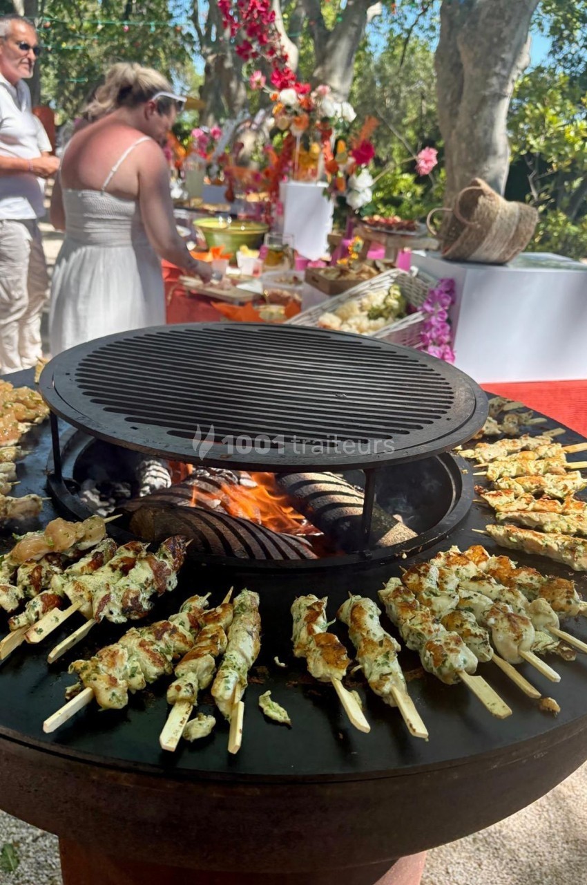 Brochettes de viande grillant sur un barbecue rond, avec une table garnie de plats et des personnes en arrière-plan.