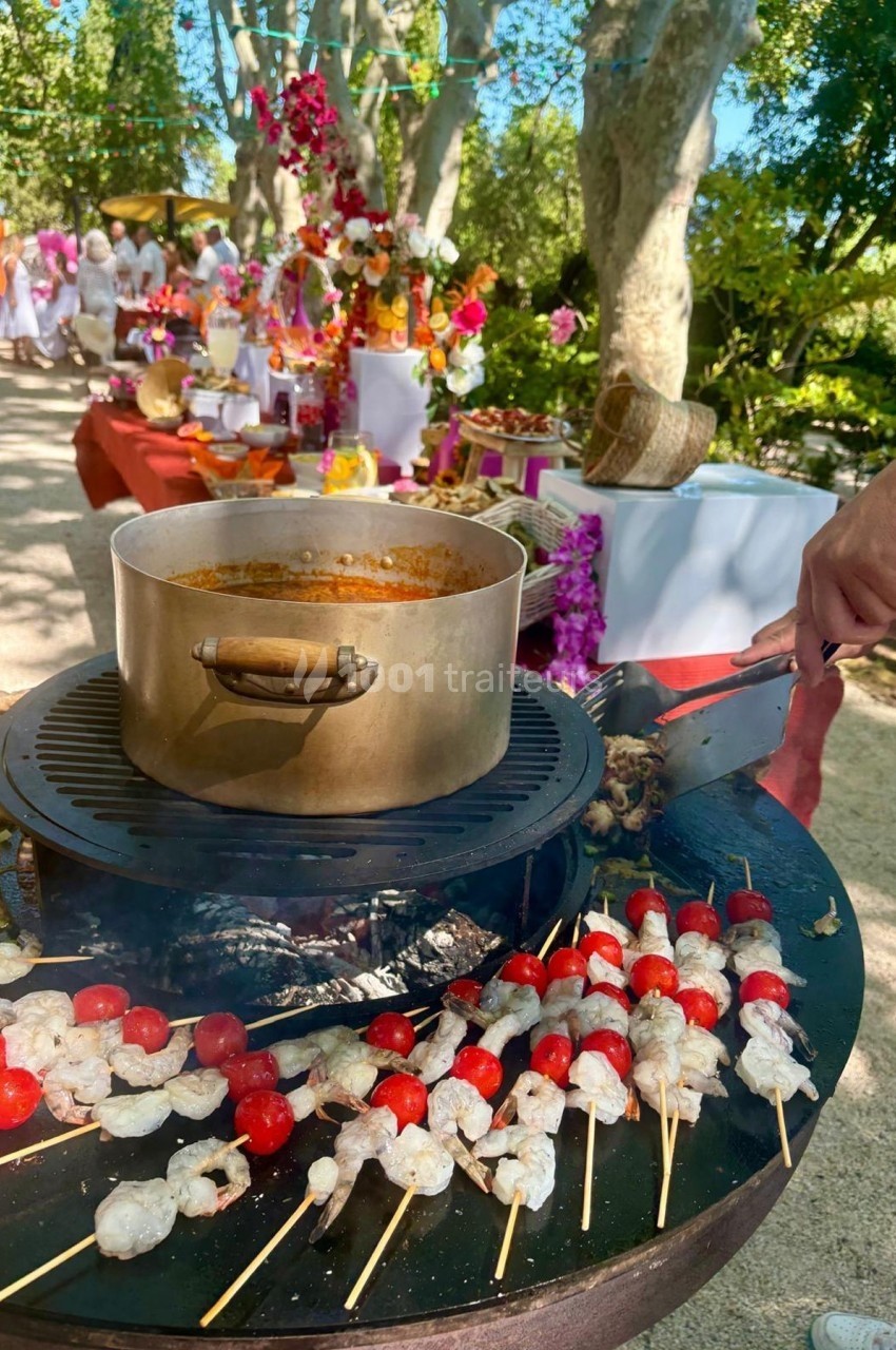 Brochettes de crevettes et tomates cerises grillées sur un barbecue rond, avec une marmite en arrière-plan dans un jardin.