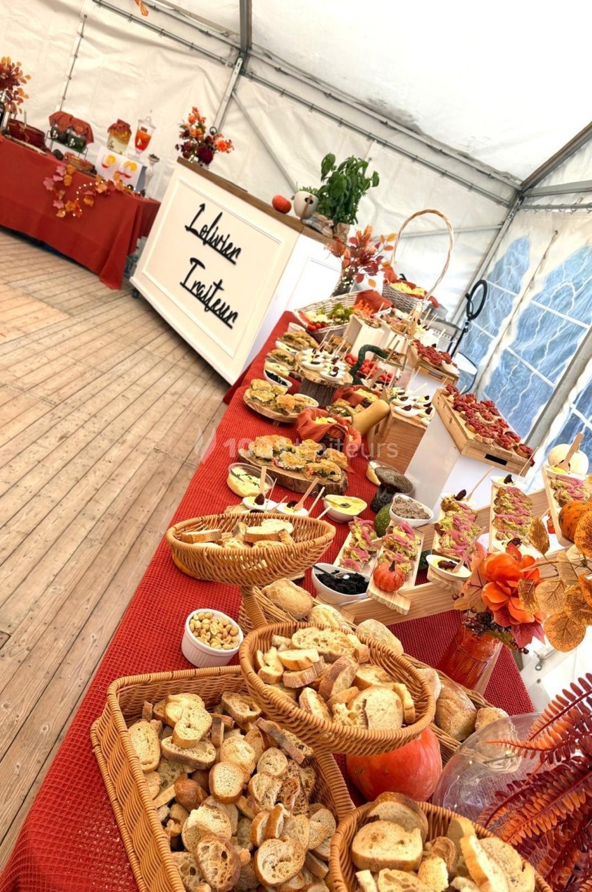 Buffet automnal sous une tente, avec pains, desserts, décorations orange et rouges, et un panneau ’L'atelier du Traiteur’.