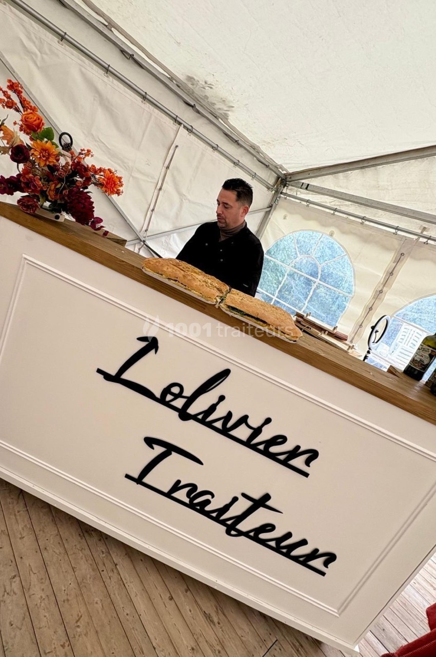 Un traiteur prépare des plats derrière un comptoir blanc décoré de fleurs dans une tente lumineuse.