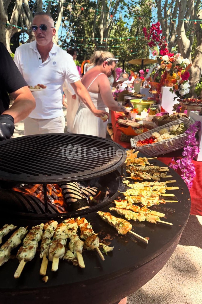 Grill circulaire avec brochettes en cuisson, buffet coloré en arrière-plan et personnes se servant de la nourriture.