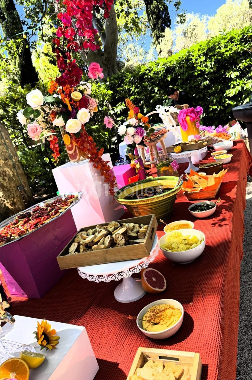 Table garnie de plats variés, fleurs colorées et décorations, disposée en extérieur sous un ciel ensoleillé.