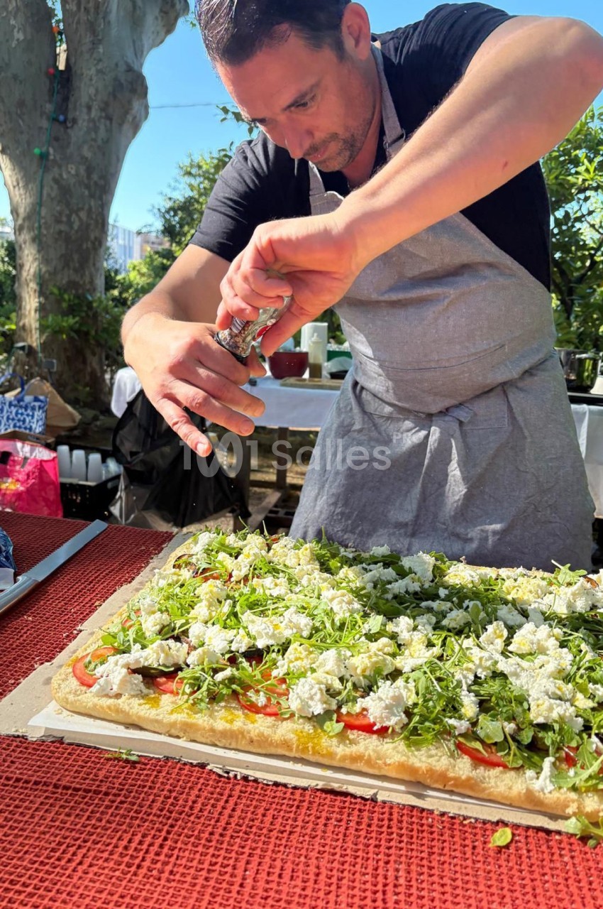 Un homme en tablier assaisonne une grande tarte salée garnie de légumes et fromage, dans un jardin ensoleillé.