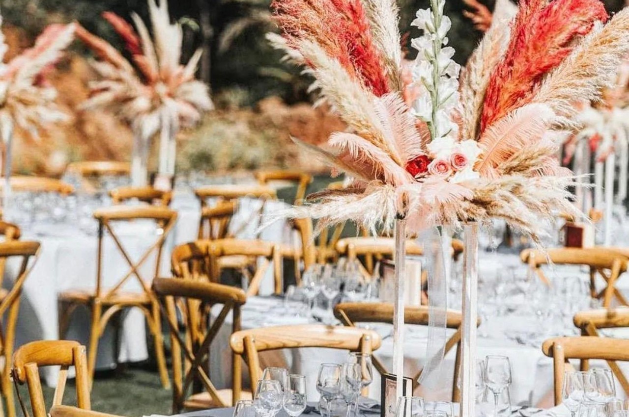 Tables de réception en extérieur décorées de nappes blanches, verres, et centres de table avec fleurs et pampas colorées.