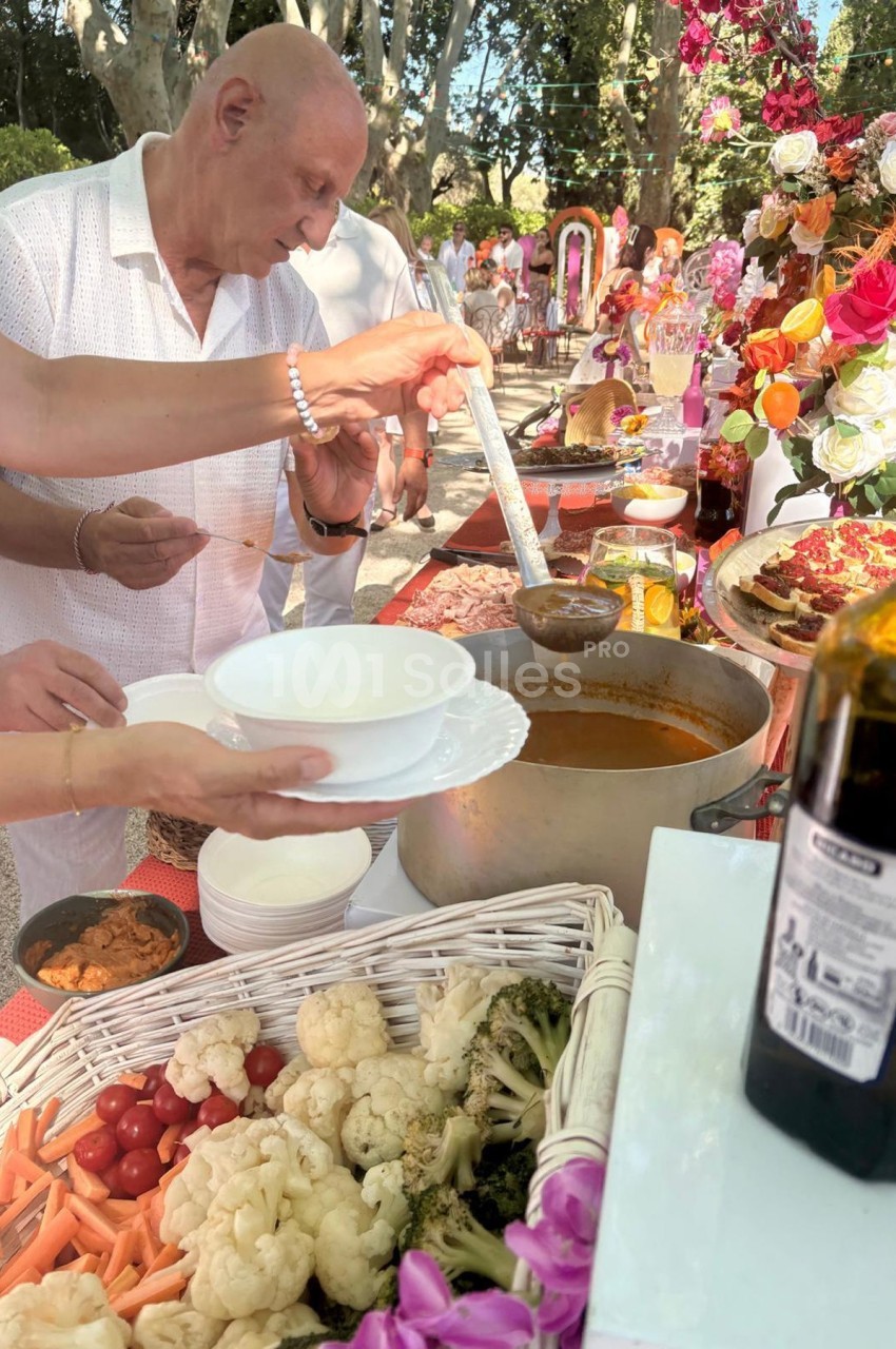 Un homme sert de la soupe dans un bol lors d'un buffet en plein air, entouré de légumes et de décorations florales.