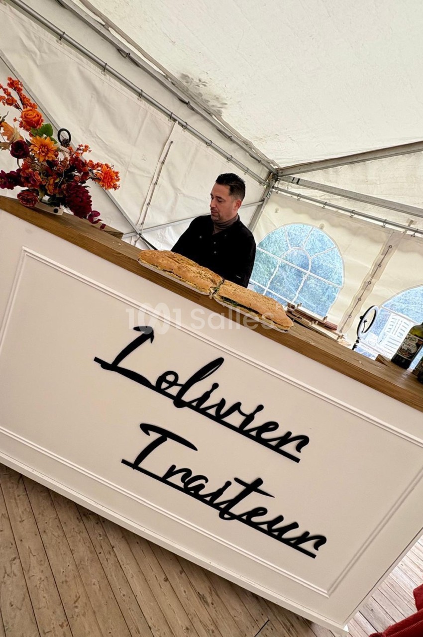 Un traiteur prépare des plats derrière un comptoir blanc décoré de fleurs dans une tente lumineuse.