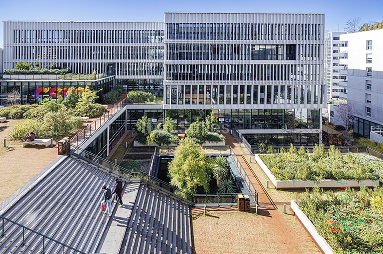 Bâtiment moderne avec façades vitrées, espaces verts et escaliers extérieurs dans un environnement urbain.