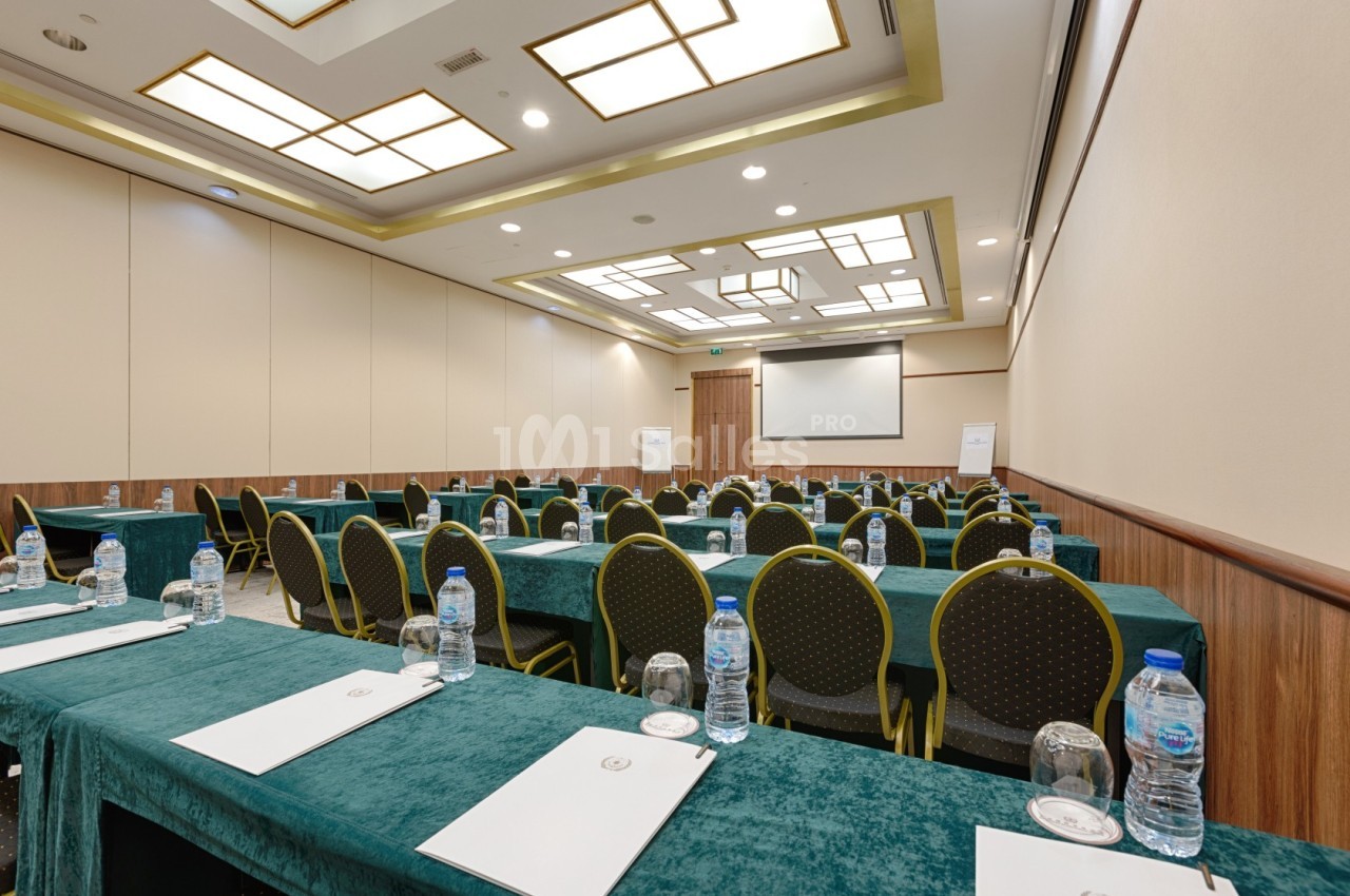 Salle de conférence avec des rangées de tables recouvertes de nappes vertes, chaises alignées et écran de projection.