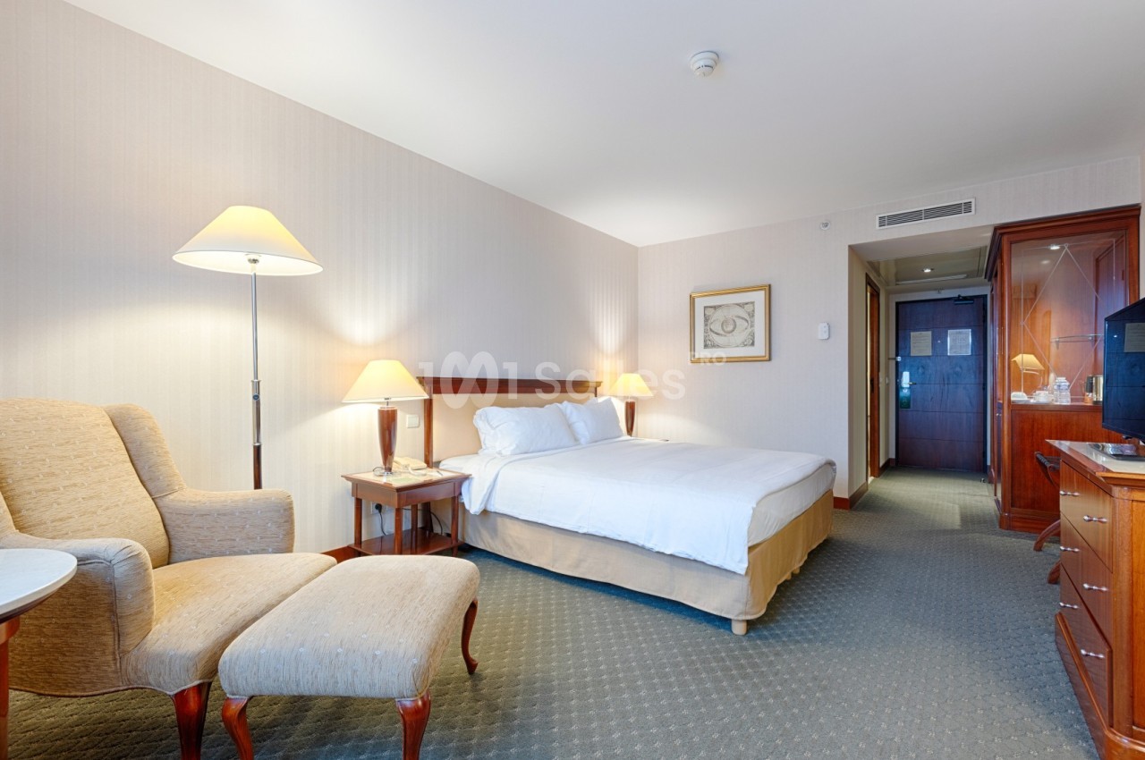Chambre d'hôtel lumineuse avec lit double, fauteuil, bureau en bois, éclairage doux et porte menant à un couloir.