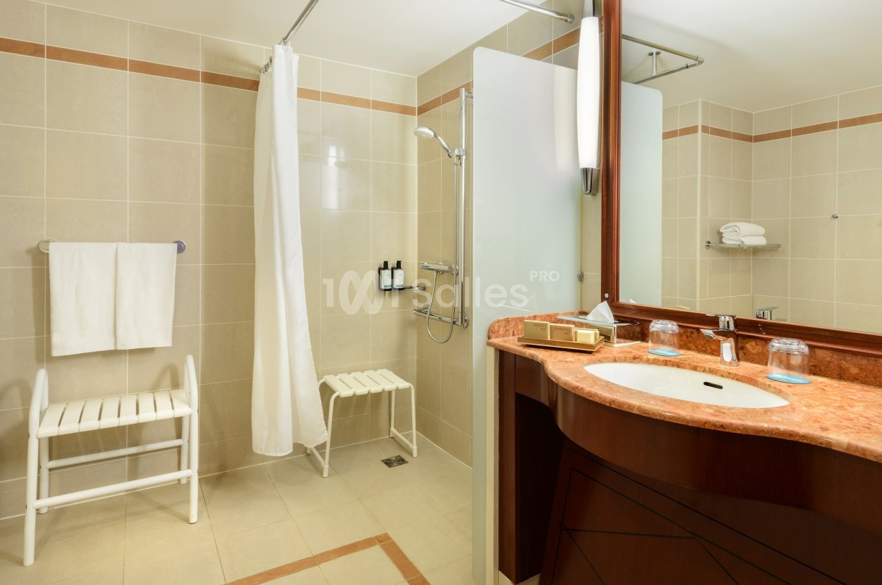 Salle de bain accessible avec douche à l'italienne, rideau, lavabo en marbre et tabourets blancs.