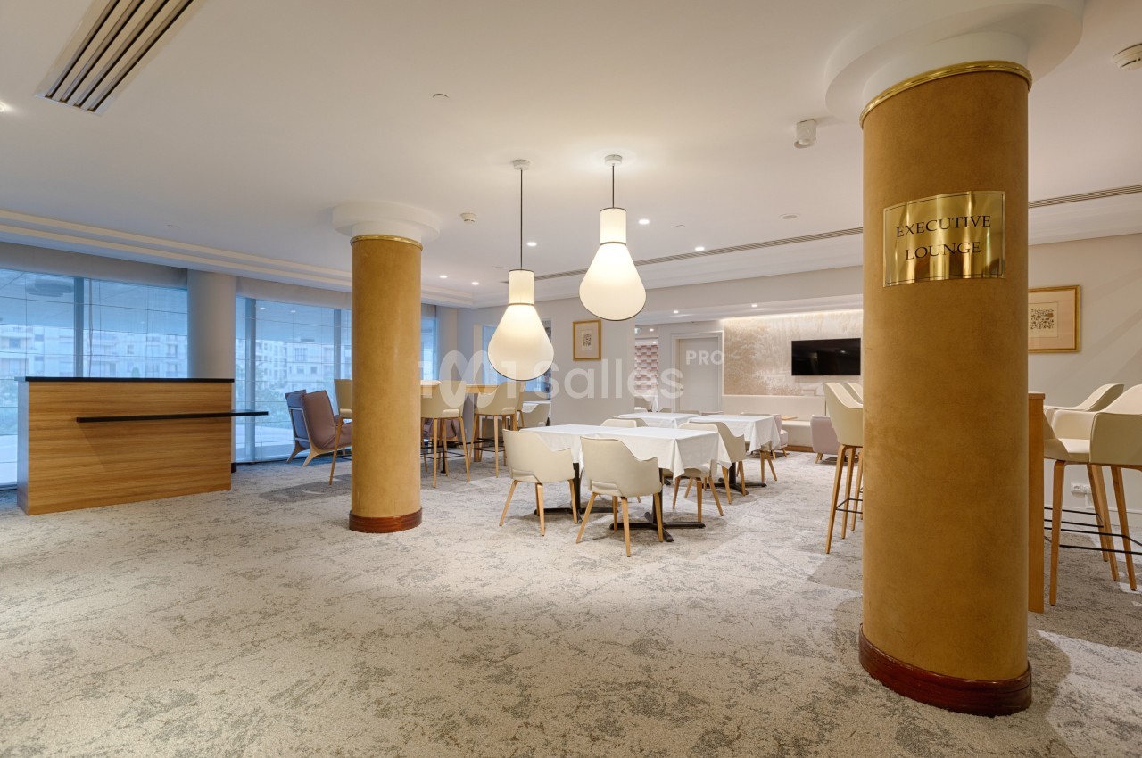 Salle lumineuse avec tables, chaises blanches, colonnes dorées et un comptoir en bois, ambiance moderne et épurée.