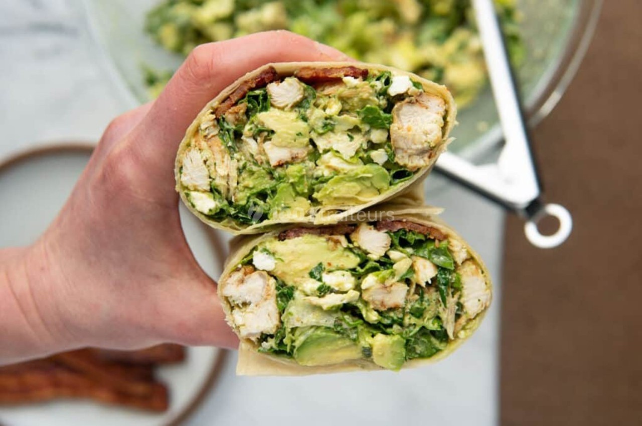 Wrap coupé en deux, garni de poulet, avocat, salade et autres ingrédients frais, tenu dans une main.