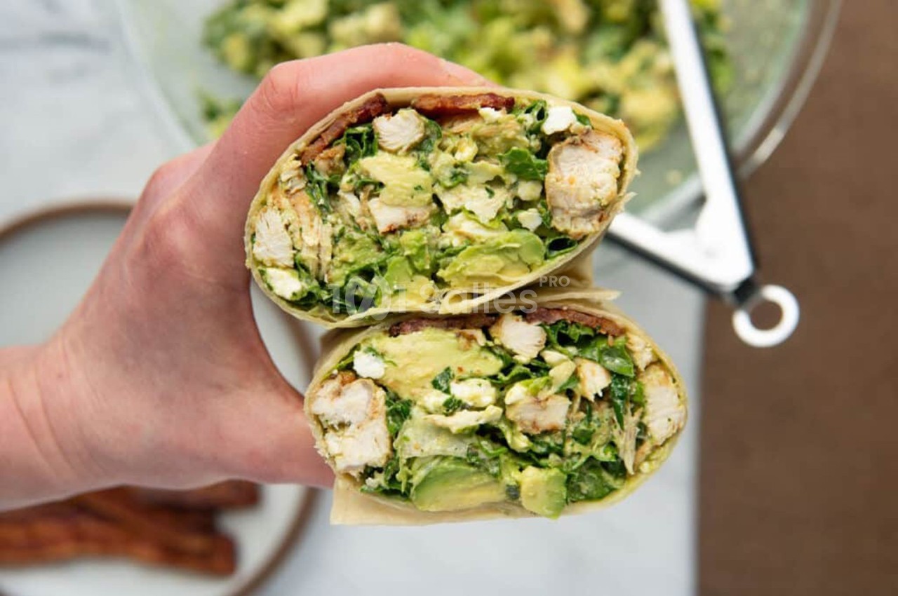 Deux wraps coupés en deux, garnis de poulet, avocat, salade et autres ingrédients frais, tenus dans une main.