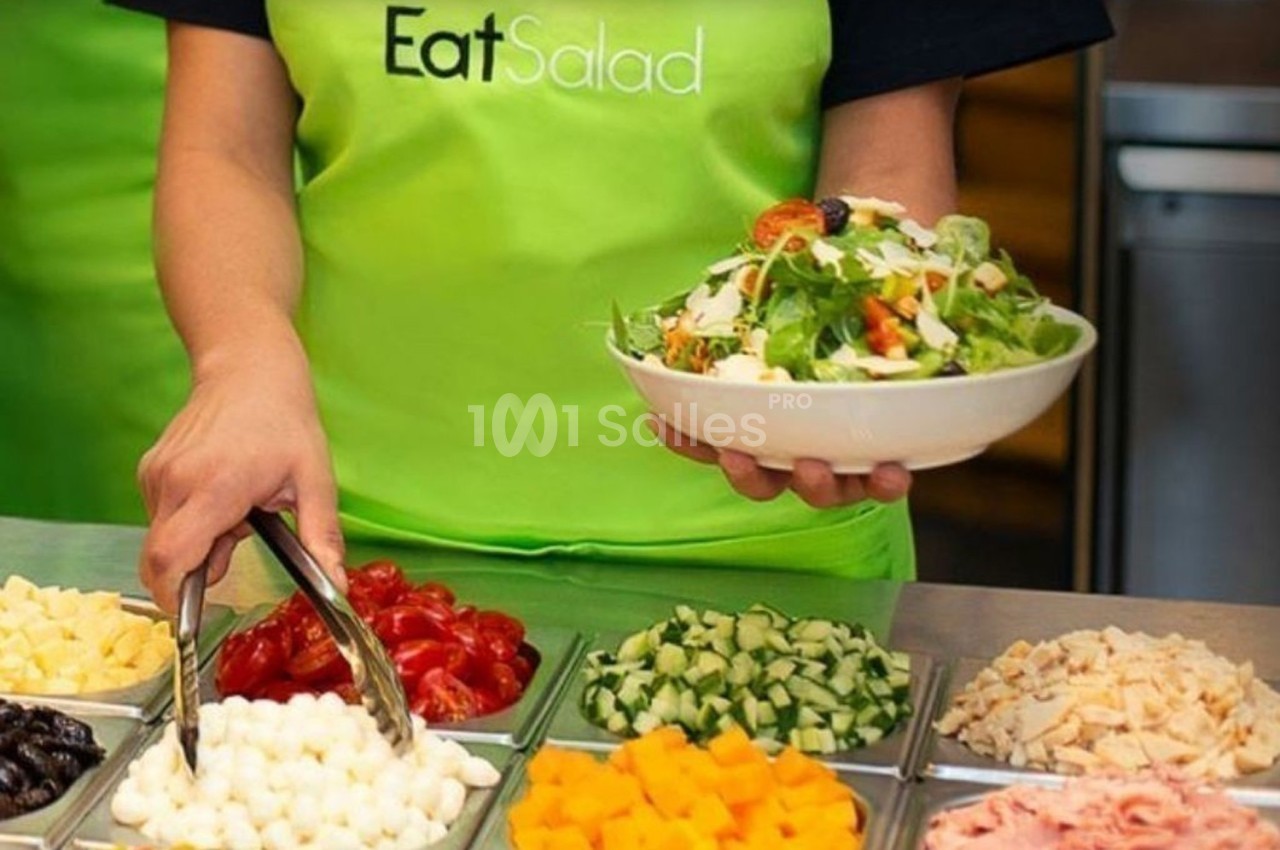Une personne sert une salade dans un bol, devant un comptoir garni d'ingrédients variés.