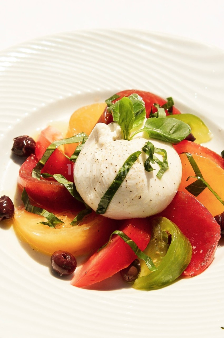 Assiette de burrata entourée de tranches de tomates colorées, olives noires et basilic frais.