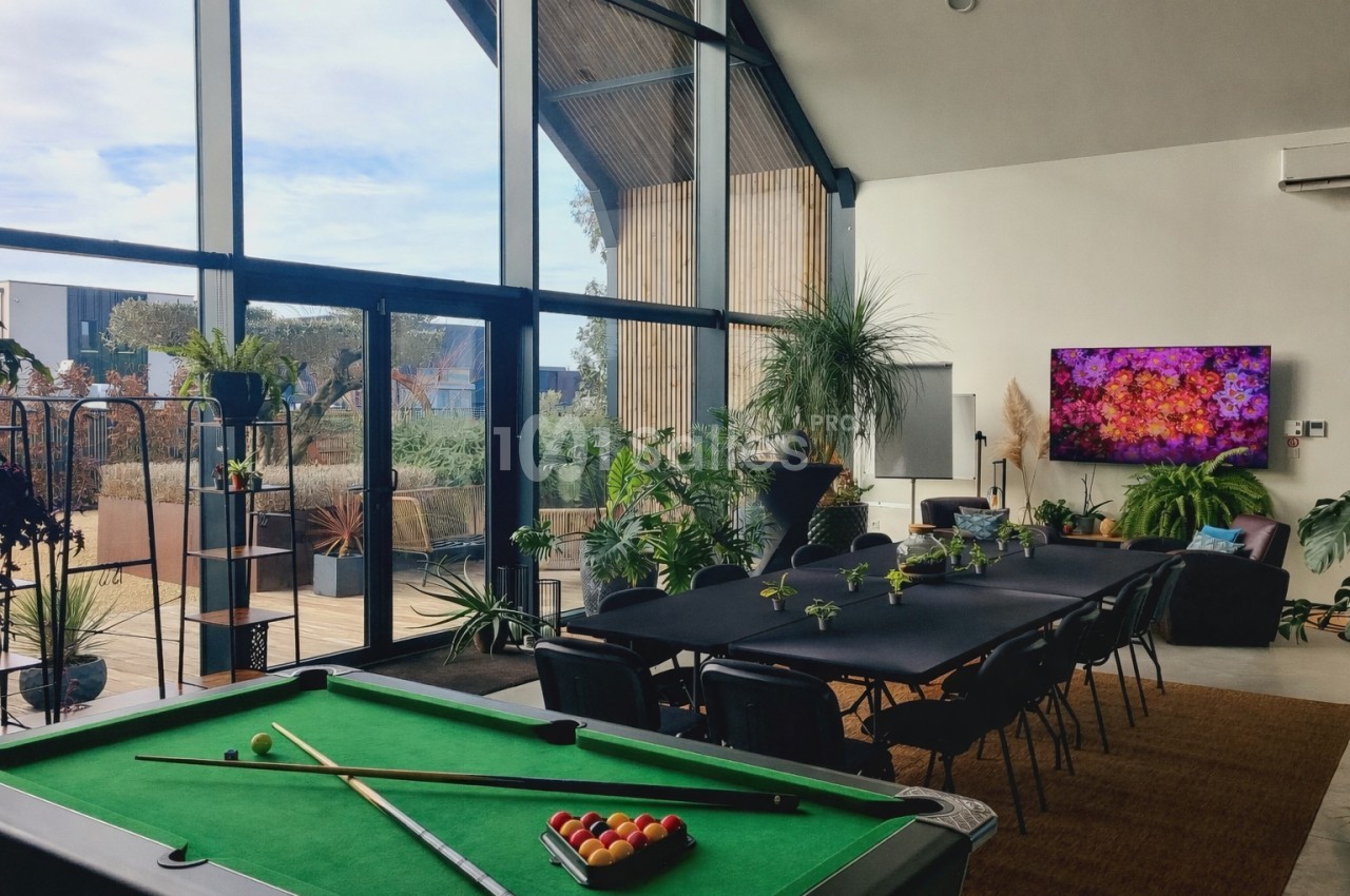 Salle lumineuse avec table de billard, grande table de réunion, plantes et baie vitrée donnant sur une terrasse.